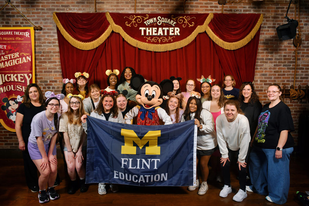 A group of people poses together in front of a brick wall with a red curtain, holding a blue flag that reads "M FLINT EDUCATION." Mickey Mouse stands in the center, wearing a colorful outfit.