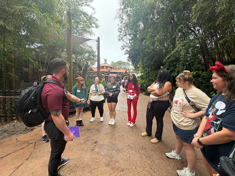 A group of people stands on a path in a lush, green area, listening to a person in a maroon shirt and backpack who is speaking to them.