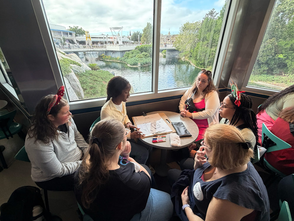 A group of people sitting around a table inside a building, with a view of a river and greenery outside the window. There are papers and markers on the table.