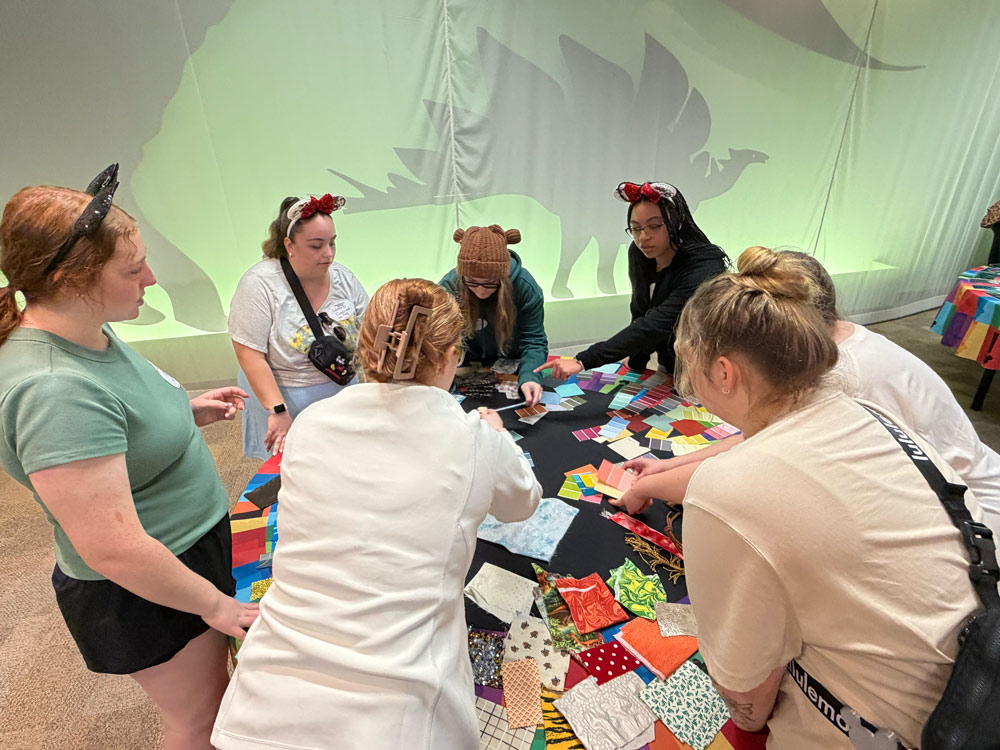 A group of people gathered around a table covered with colorful fabric swatches, discussing and selecting materials. A large silhouette of a dinosaur is visible in the background.