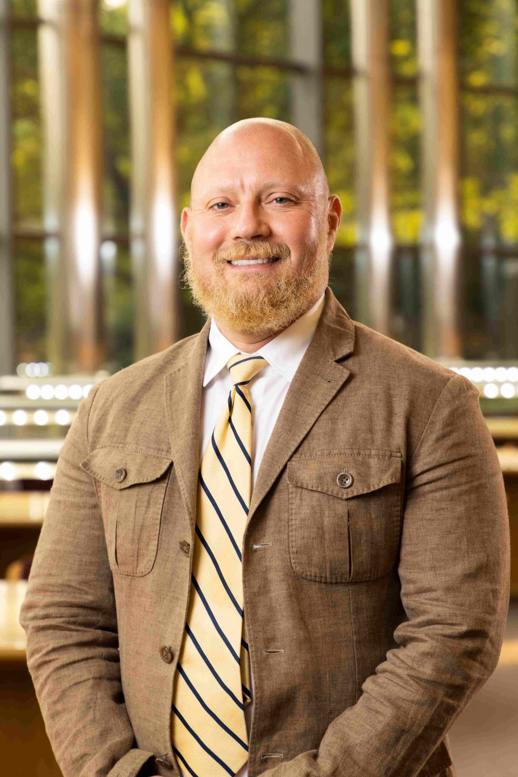A professional portrait of a smiling man with a bald head and blonde beard. He is wearing a white dress shirt, yellow tie with navy-blue stripes, and a tan, textured blazer with pocket flaps. He is standing in a sunlit room with a blurred background of large windows and greenery.