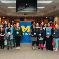 A group of people stands together in a room, with a blue banner featuring a yellow "M" for the University of Michigan-Flint in the background. Some individuals are wearing name tags. The setting appears to be a conference or meeting space.
