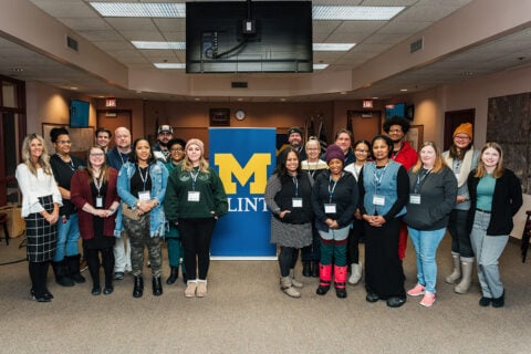 A group of people stands together in a room, with a blue banner featuring a yellow "M" for the University of Michigan-Flint in the background. Some individuals are wearing name tags. The setting appears to be a conference or meeting space.