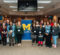 A group of people stands together in a room, with a blue banner featuring a yellow "M" for the University of Michigan-Flint in the background. Some individuals are wearing name tags. The setting appears to be a conference or meeting space.