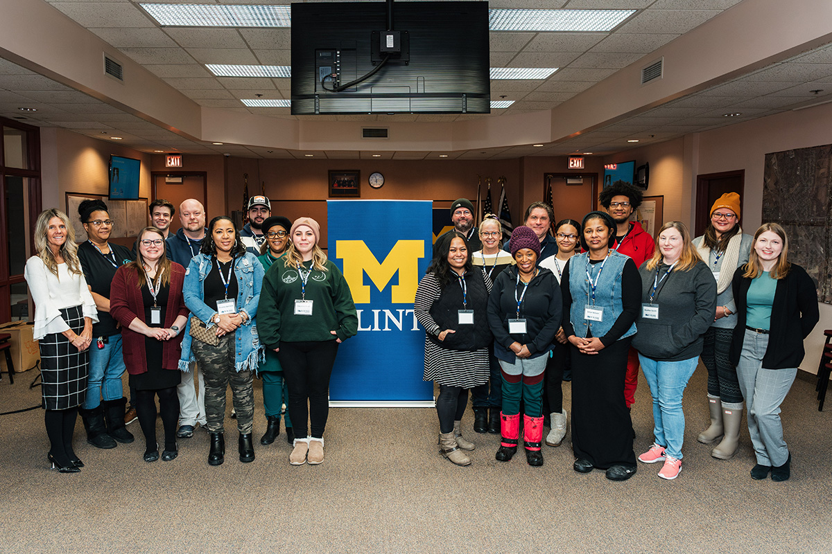 A group of people stands together in a room, with a blue banner featuring a yellow "M" for the University of Michigan-Flint in the background. Some individuals are wearing name tags. The setting appears to be a conference or meeting space.