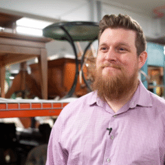 A person wearing a checkered shirt stands in an antique store, with wooden furniture and shelves in the background.