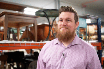 A person wearing a checkered shirt stands in an antique store, with wooden furniture and shelves in the background.