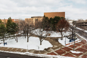 A winter scene showing UM-Flint's campus with snow-covered ground, trees, and buildings in the background. A street sign and a parking structure are visible on the right.
