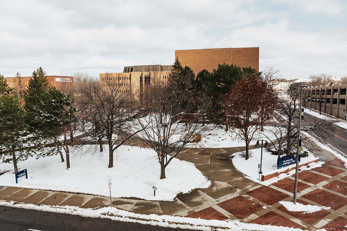 A winter scene showing UM-Flint's campus with snow-covered ground, trees, and buildings in the background. A street sign and a parking structure are visible on the right.
