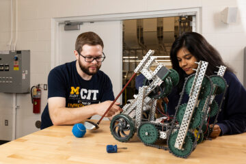 Two individuals are working together on a robotic project, focusing on a complex robot made of metal parts and wheels on a wooden table. Tools and a blue ball are also visible in the scene.