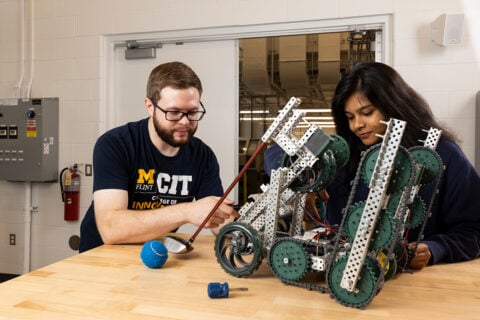 Two individuals are working together on a robotic project, focusing on a complex robot made of metal parts and wheels on a wooden table. Tools and a blue ball are also visible in the scene.