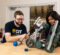 Two individuals are working together on a robotic project, focusing on a complex robot made of metal parts and wheels on a wooden table. Tools and a blue ball are also visible in the scene.