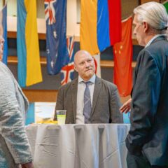 A group of people are engaged in conversation around a high-top table. Behind them, various international flags hang from an atrium's ceiling.
