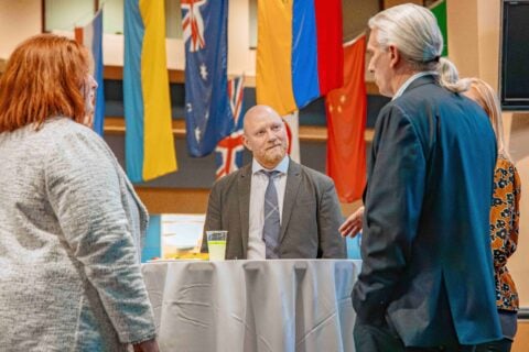 A group of people are engaged in conversation around a high-top table. Behind them, various international flags hang from an atrium's ceiling.