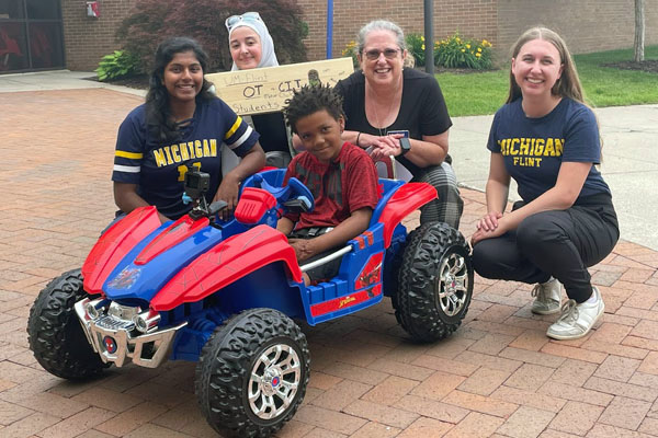 Shalini Augenstein (far left), a 2024 OTD graduate, Marwa Hammami (left), a 2026 cybersecurity graduate, assistant professor Donna Case (center) and Elizabeth Mansfield (right), a 2024 OTD graduate were part of a group that adapted a Power Wheels car for Flint child Kaden Stevenson (front).