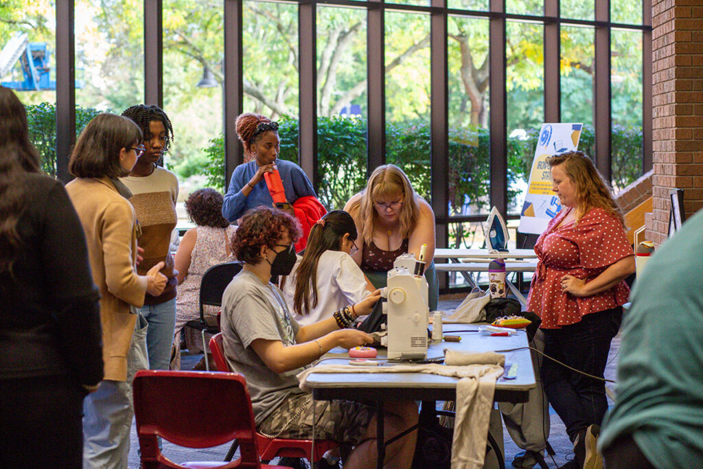 A group of people is gathered around a table with a sewing machine, fabric, and various sewing supplies in a well-lit indoor space with large windows.