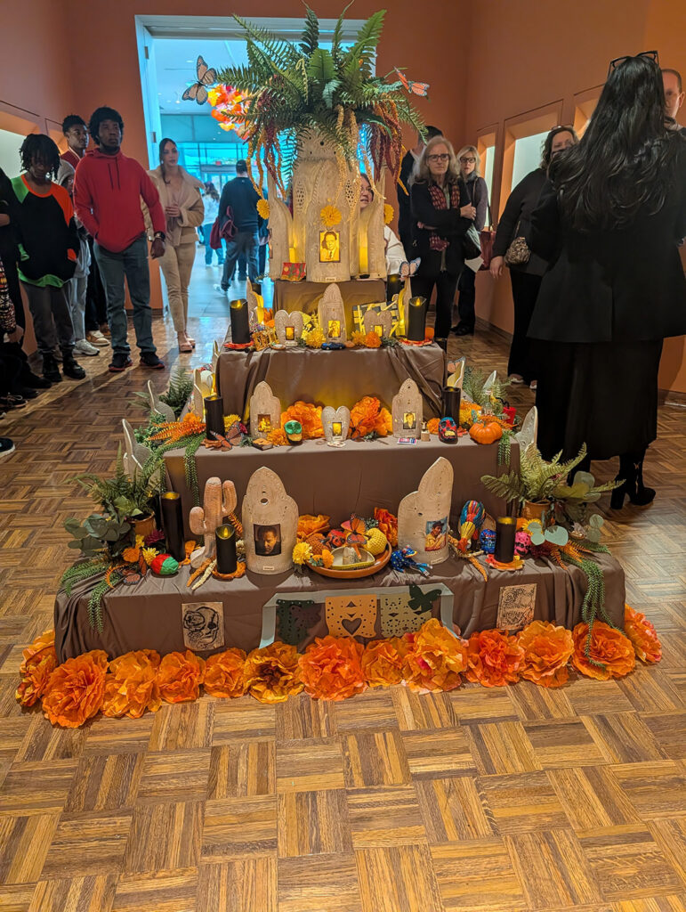 A colorful altar decorated with marigolds, candles, and various offerings, surrounded by people in a gallery setting.