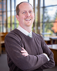 A person in a brown sweater with arms crossed, standing in a well-lit room with large windows and wooden furniture in the background.