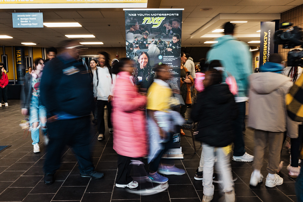 FLINT_Revving Up STEM 2026_0002 A busy indoor space with people walking past a promotional banner for NXG Youth Motorsports.