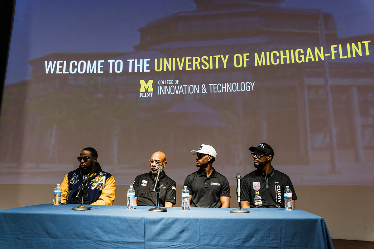FLINT_Revving Up STEM 2026_0007 A panel of four individuals seated at a table with water bottles, in front of a screen displaying "Welcome to the University of Michigan-Flint, College of Innovation & Technology."