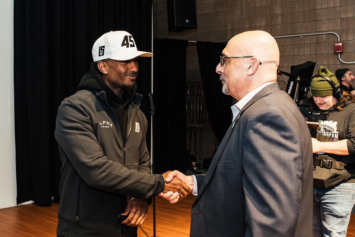 FLINT_Revving Up STEM 2026_0024 Two men are shaking hands in a well-lit indoor setting. One man is wearing a black jacket and a white cap, while the other is in a suit.