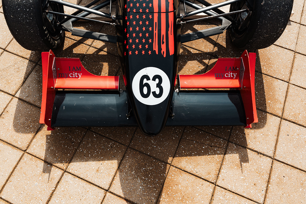 FLINT_Revving Up STEM 2026_0032 Top view of a race car featuring a black body with red accents, number 63, and the text "I AM my city" on the side wings, parked on a patterned surface.