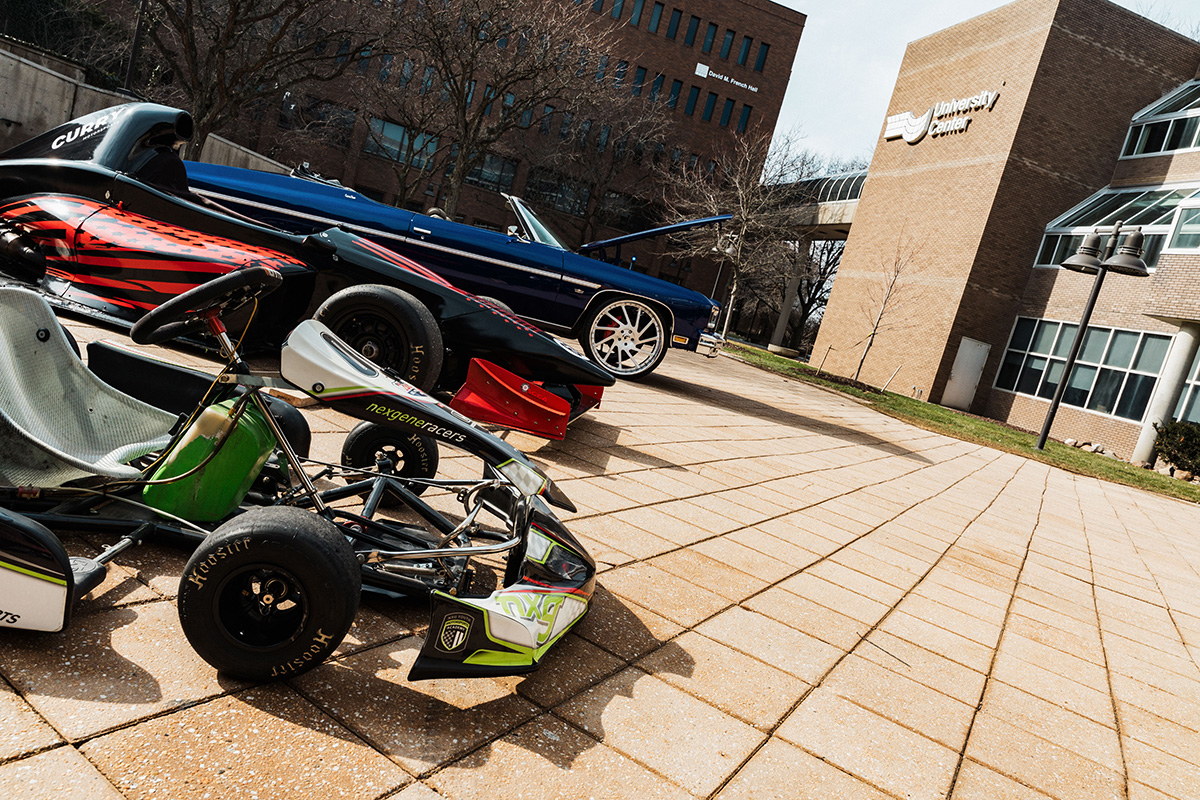 A go-kart and two race cars are parked on a paved area in front of a university building, with trees in the background.