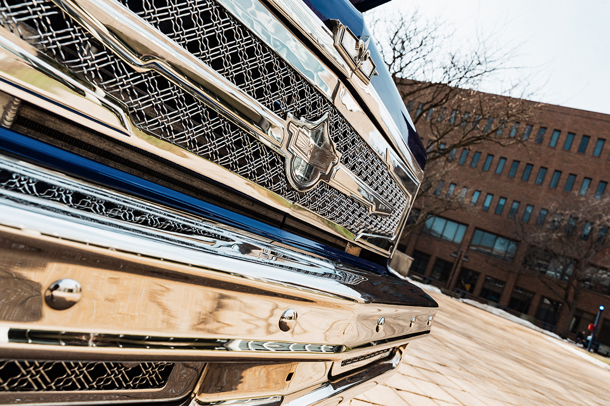 FLINT_Revving Up STEM 2026_0035 Close-up of a shiny chrome front grille of a truck, featuring a distinctive emblem and intricate mesh design, with a blurred background of buildings and trees.