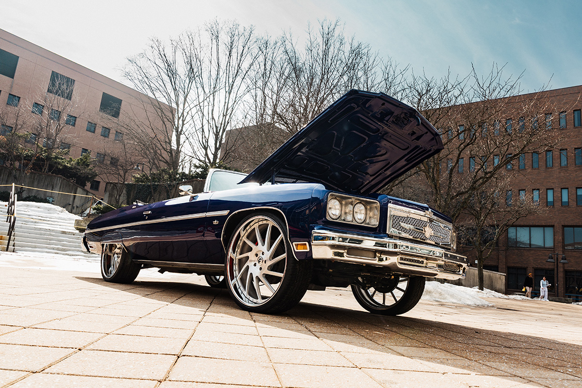 FLINT_Revving Up STEM 2026_0036 A classic blue convertible car with its hood open, parked on a brick surface, with trees and buildings in the background.