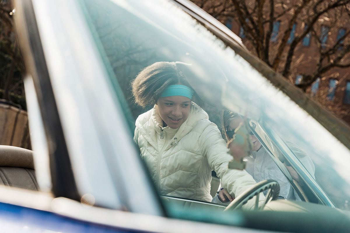 FLINT_Revving Up STEM 2026_0048 A person in a white jacket is leaning into a car, with another person partially visible in the background. The scene is viewed through the car's windshield.