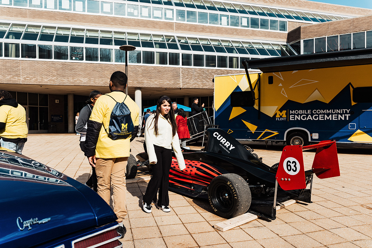 FLINT_Revving Up STEM 2026_0051 A group of people is gathered outside a building, with a black race car featuring red accents and the number 63 in the foreground. A blue vehicle with "MOBILE COMMUNITY ENGAGEMENT" is parked nearby. Some individuals are wearing yellow shirts and a backpack with a university logo.