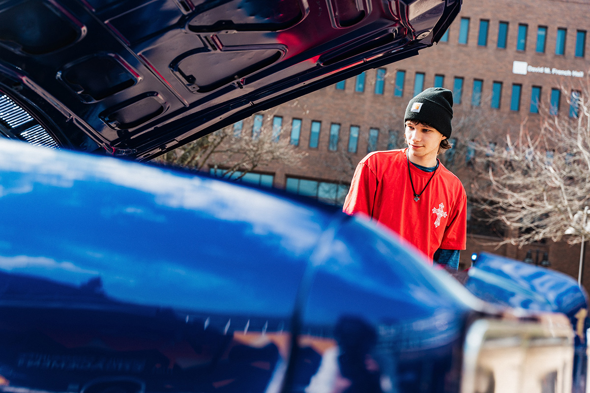 FLINT_Revving Up STEM 2026_0060 A person in a red shirt and black beanie stands near the open hood of a blue car, with a building and trees in the background.