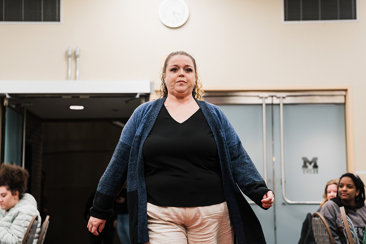 A person walks confidently down a runway, wearing a black top and a blue cardigan, with light-colored pants. In the background, there are people seated and a clock on the wall.