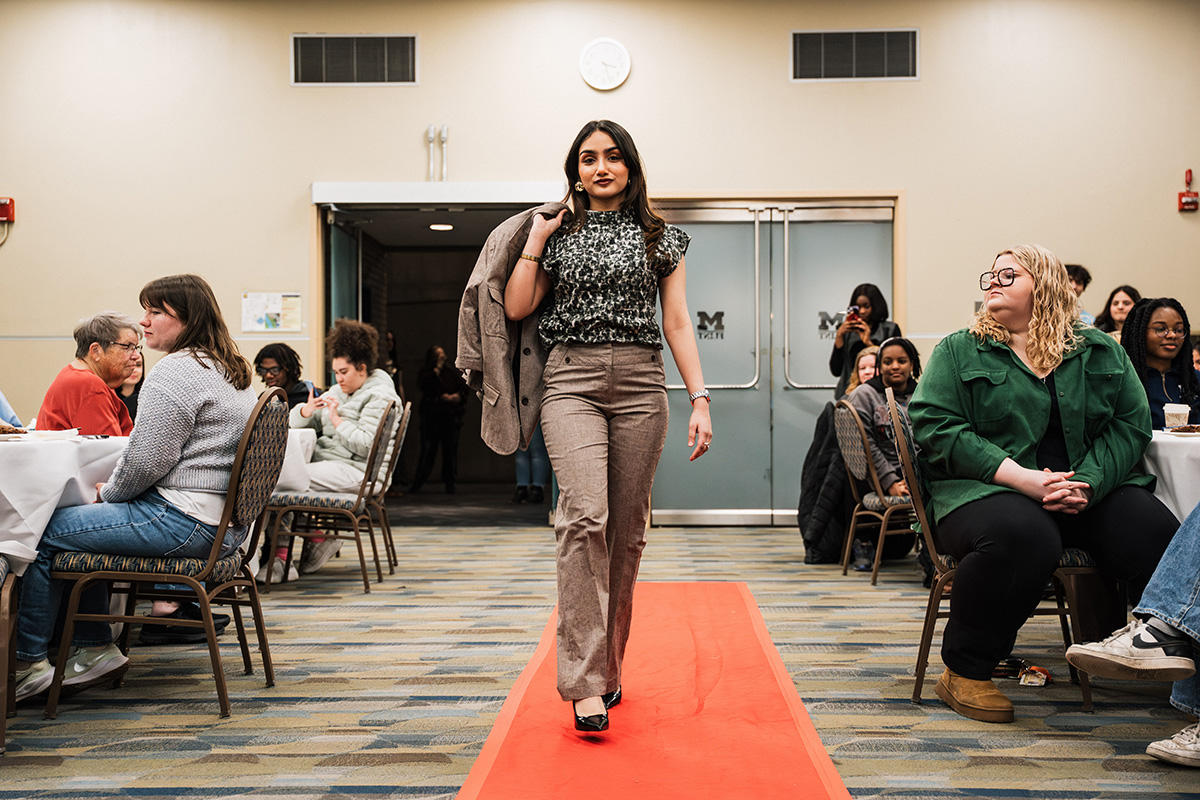 A person walks down a red carpet in a room filled with seated guests at tables, wearing a patterned top and beige pants, holding a jacket.