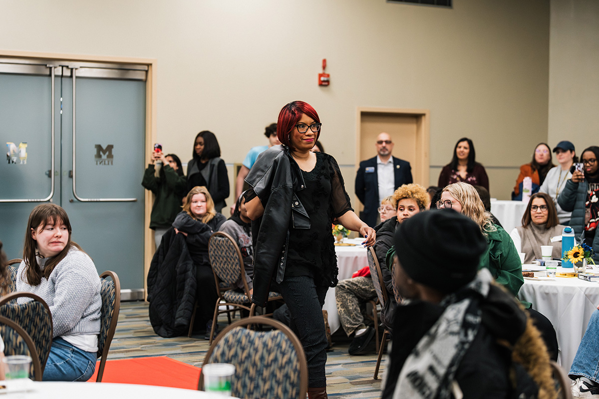 A person with red hair walks through a room filled with seated individuals at tables.