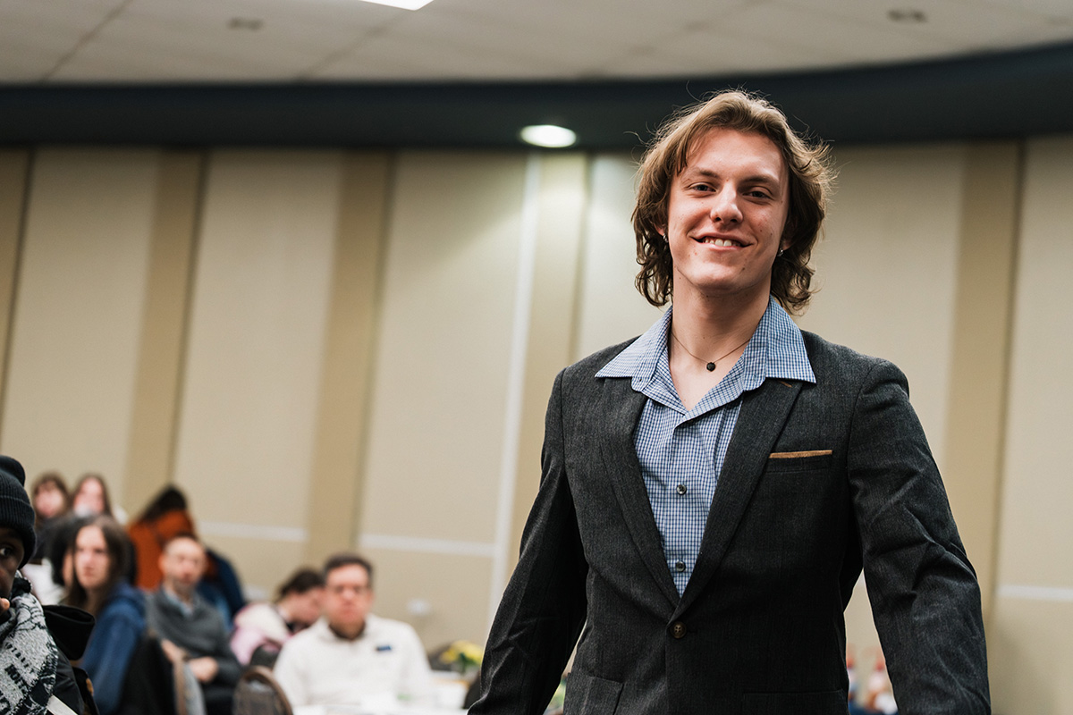 A person wearing a dark suit jacket over a light blue checkered shirt walks confidently in a room filled with seated attendees.