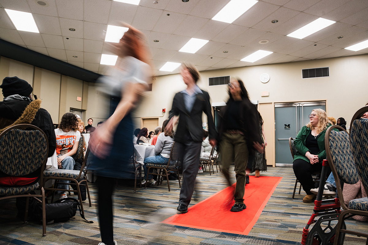 A fashion show taking place indoors, with models walking on a red carpet in front of an audience seated at tables.