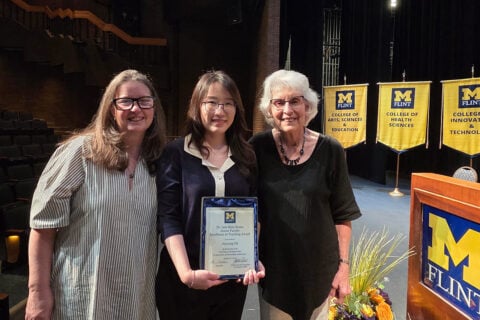 Three individuals stand together on a stage, holding a framed award certificate. The backdrop features banners for the University of Michigan-Flint's College of Arts, Sciences & Education, College of Health Sciences, and College of Innovation & Technology.