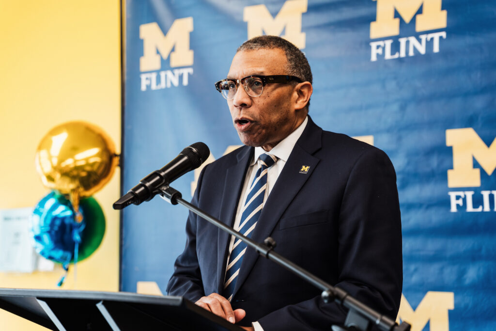 suit and tie speaking at a podium with a UM-Flint backdrop.