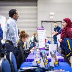 A group of people interacting at a career fair, with a table displaying informational materials and a sign listing core values.