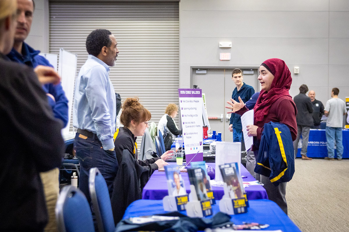 A group of people interacting at a career fair, with a table displaying informational materials and a sign listing core values.