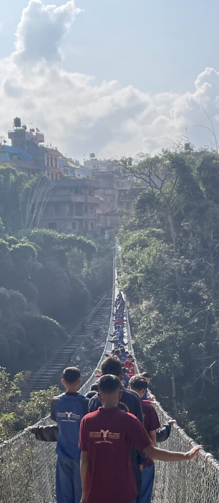 A group of people walking across a suspension bridge surrounded by greenery and buildings in the background under a cloudy sky.
