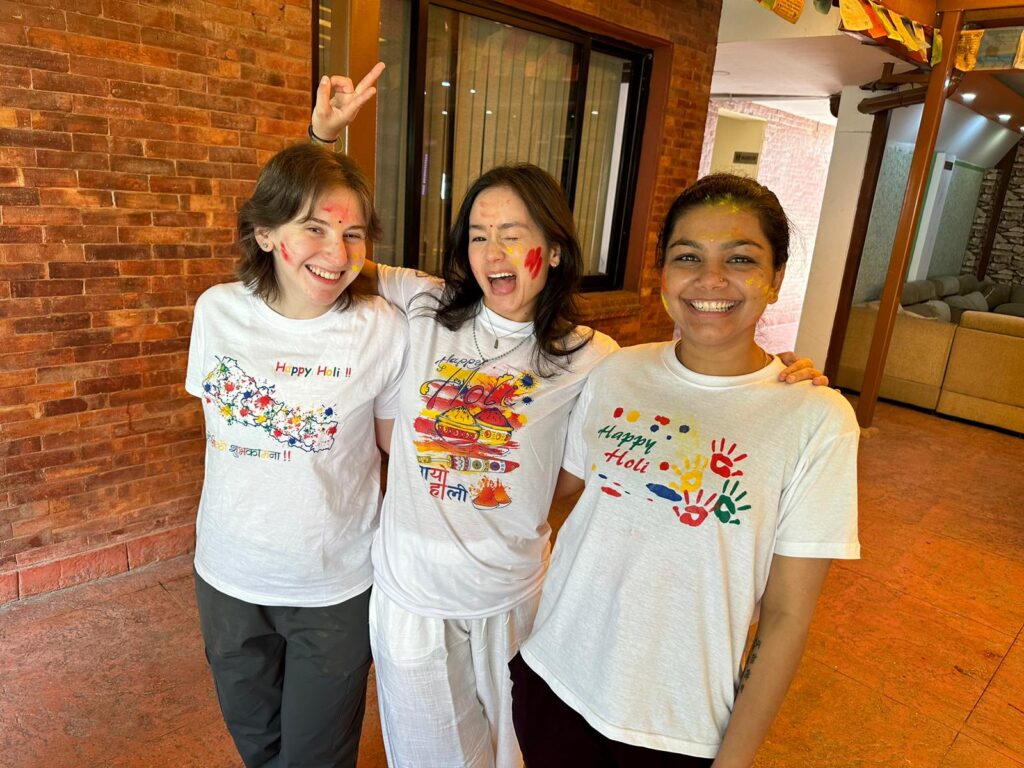 Three individuals are standing together, wearing white t-shirts with colorful designs celebrating "Happy Holi." The background features a brick wall and a glimpse of a room with furniture.