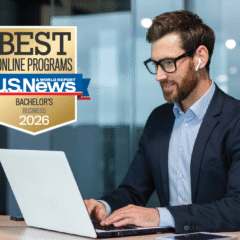 A man wearing glasses, a navy-blue suit and wireless earbuds is sitting at a desk with a laptop. A golden badge is in the upper left-hand corner belonging to U.S. News and World Report. The badge reads "Best Online Programs: Bachelor's Business 2026."