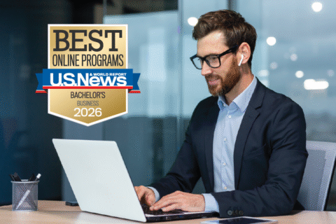 A man wearing glasses, a navy-blue suit and wireless earbuds is sitting at a desk with a laptop. A golden badge is in the upper left-hand corner belonging to U.S. News and World Report. The badge reads "Best Online Programs: Bachelor's Business 2026."