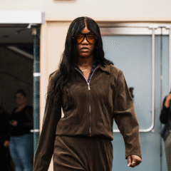 A person walks confidently in a brown matching outfit, with a zip-up jacket and pants, in a public space. In the background, a few people are watching and taking photos.