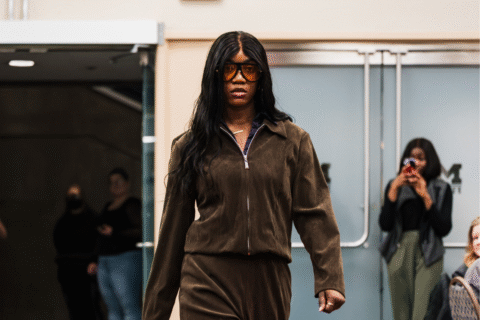 A person walks confidently in a brown matching outfit, with a zip-up jacket and pants, in a public space. In the background, a few people are watching and taking photos.