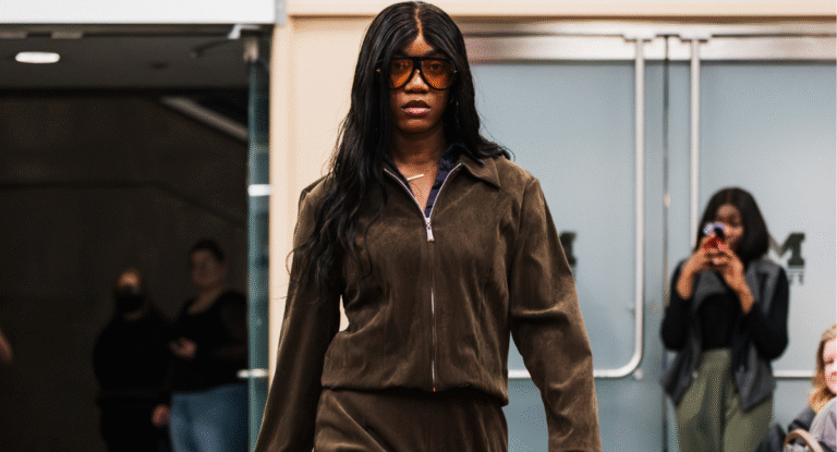 A person walks confidently in a brown matching outfit, with a zip-up jacket and pants, in a public space. In the background, a few people are watching and taking photos.