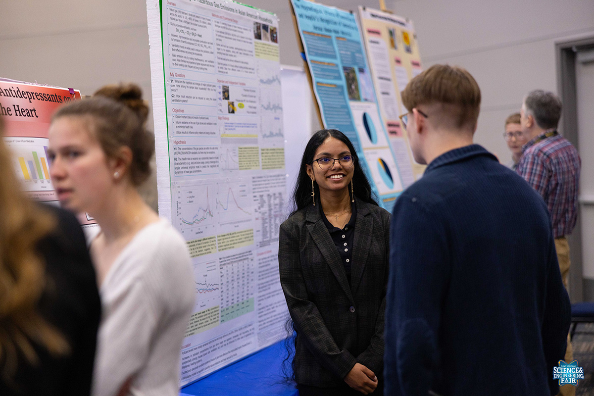 A group of people is engaged in discussion at a science fair, with several research posters displayed in the background.