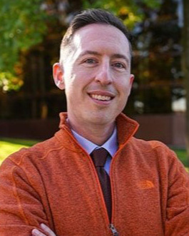 A person wearing an orange jacket and a tie stands outdoors with arms crossed, surrounded by greenery.